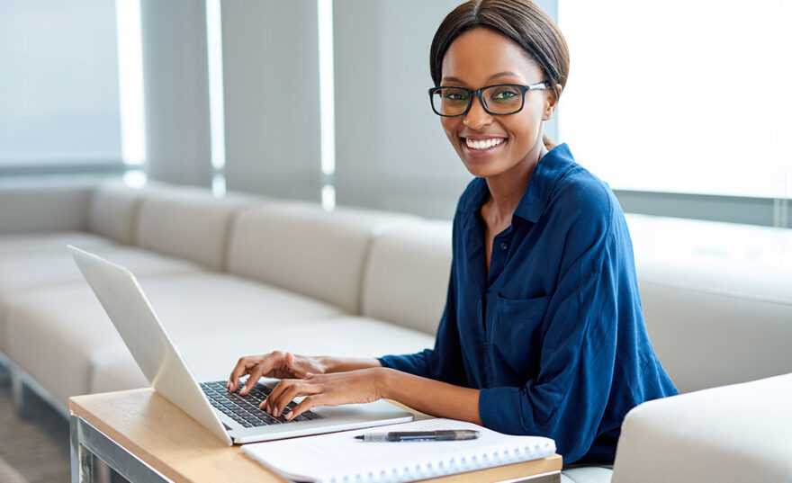 Portrait of a smiling young woman wearing glasses working on a laptop while sitting at a table in a modern office